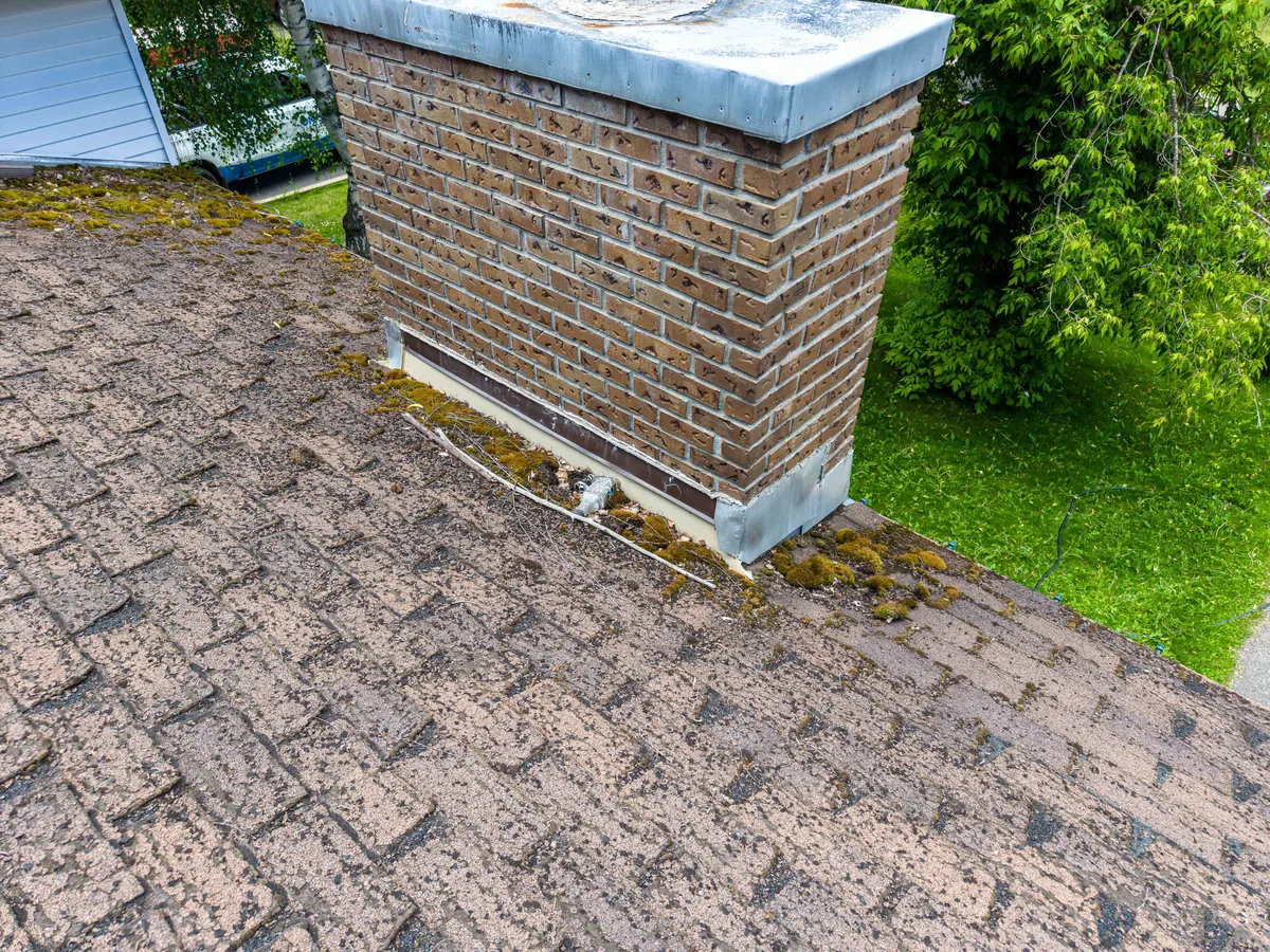 Close aerial view of a brick chimney, adjacent shingles, flashing, and moss